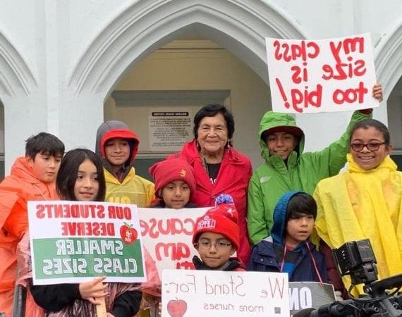 学生们和穿着雨衣的德洛丽丝·韦尔塔站在一起，举着“为Ed而红”的牌子